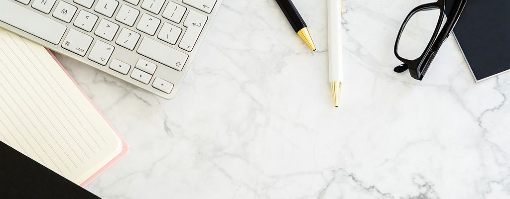 Mesa branca com teclado de computador a esquerda junto com caderno ; canetas branca e preta no centro superior da imagem e um óculo com um caderno preto a direita