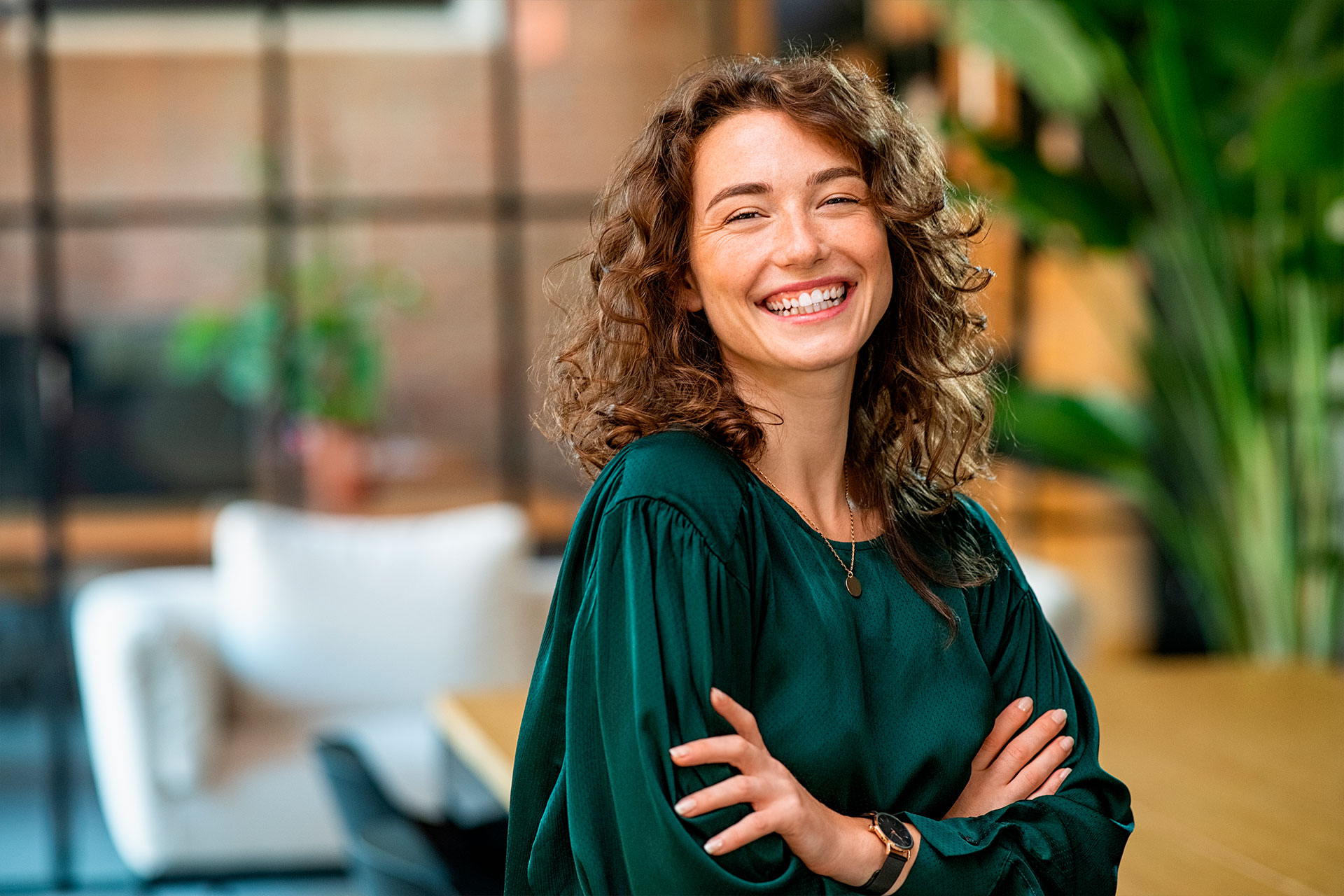 mulher branca de cabelo ondulado com braços cruzados, blusa verde escura de manga comprida sorrindo