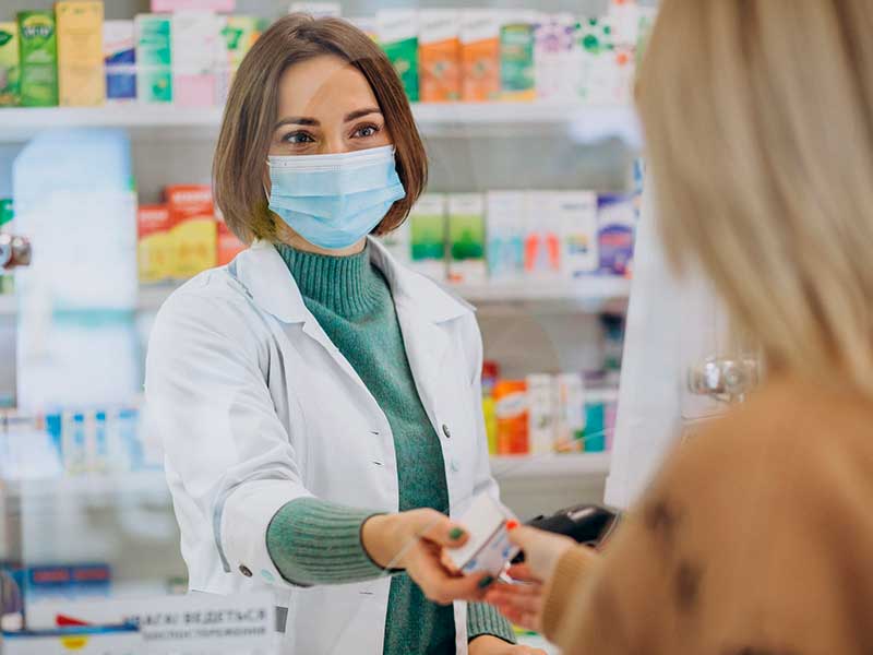descontos-em-farmacias farmacêutica de jaleco entregando um remédio para uma cliente de costas
